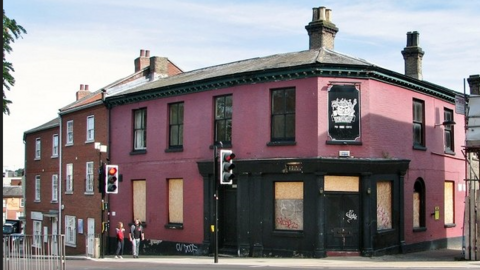 A boarded-up two-storey building, viewed from across the street. It is painted a deep rose pink with black-framed sash windows on the upper floor. The ground floor windows are all covered in chipboard. The old entrance is on the corner of the building, at an angle, and features two black doors and a wooden black frame surrounding the door and two boarded-up windows either side. There is illegible graffiti on this chipboard.