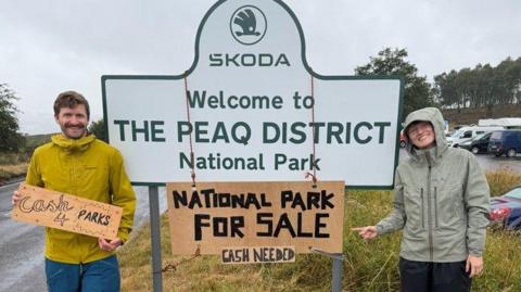 Two people at the side of a road sign which reads SKODA WELCOME TO THE PEAQ DISTRICT NATIONAL PARK. A cardboard sign is hung around the larger sign which reads NATIONAL PARK FOR SALE CASH NEEDED.