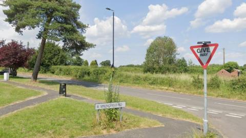 A road lined with trees and bushes. Another road, also lined with greenery, joins it at a junction.
