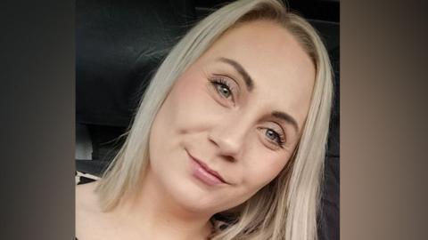 Selfie of Nicola Burns, who has shoulder-length straight blonde hair and is smiling with her mouth closed nd ger head tilted to the right. The backgroud is very dark