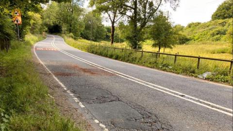 The A436 road. It is surrounded on both sides by green space and trees. The road itself is very windy and has potholes.