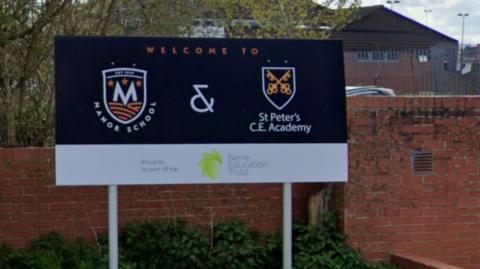 School sign at entrance for Manor School and St Peter's Academy. Both have logos on the sighn, which has a blue background and a white band at the bottom. A school building is visible behind.