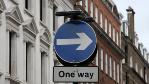 A circular blue sign with a white arrow on it with a white plate underneath that says "One way"