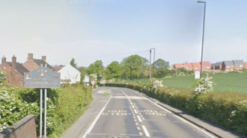 Street view at Grassmoor village sign in Derbyshire. Houses can be seen nearby on the left, with a field on the other side of the road, with other houses in the distance. It is a sunny day. 