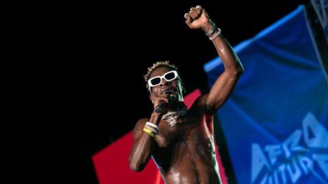 A bare-chested Shatta Wale singing at a concert. He is wearing white plastic sunglasses and has his fist raised.