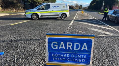 A blue Garda road closed sign sits in the middle of a road. In the background, there's a white van with Garda written on it and an officer is speaking to the driver of a grey car. It is a bright day with blue sky off in the distance.