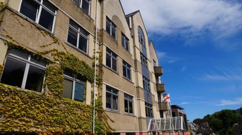 This is an image of the exterior of Frossard House. It is a building with a yellow exterior, with a flagpole bearing a Guernsey flag.