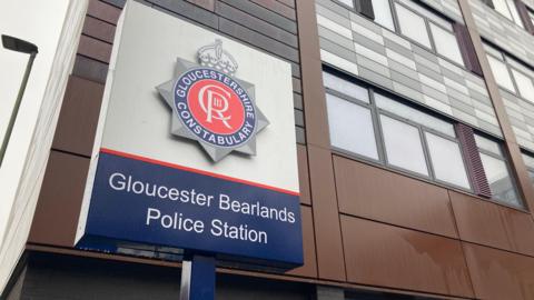 A photograph of a sign outside a Gloucestershire Police station with the constabulary's logo. underneath the logo which bears the royal insignia of King Charles III is the writing, 'Gloucester Bearlands Police Station'.