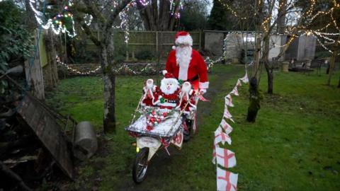 Barry Illing is dressed as Santa and wheeling a decorated wheelbarrow through a garden.