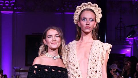 Shonagh Murray (left) with a model wearing one of her designs. Shonagh has long blonde hair which has been curled and wears a black knitted off-the-shoulder dress and a pearl Vivienne Westwood necklace. The model on the right is considerably taller than Shonagh and wears a low-cut cream knitted dress with a matching crown-style headband. The stage behind them is lit purple. 