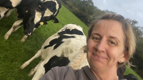 Marie Stephenson, woman with blonde hair smiling. She is in a field with several cows in the background with her.