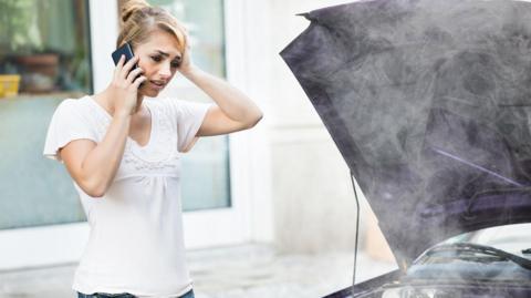 Young woman using mobile phone while looking at broken down car on street.