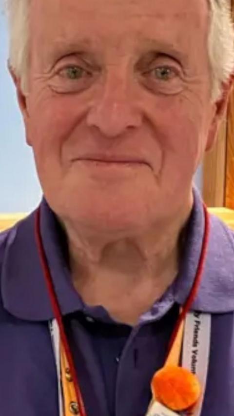 A white old man wearing a purple top with white hair smiles at the camera 