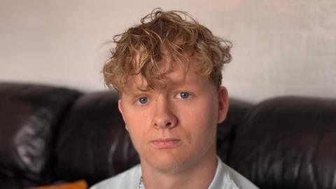 A head and shoulders of Harry Pyett, who has curly light brown hair and blue eyes, and is looking at the camera. He is sitting on a dark-coloured leather sofa indoors.