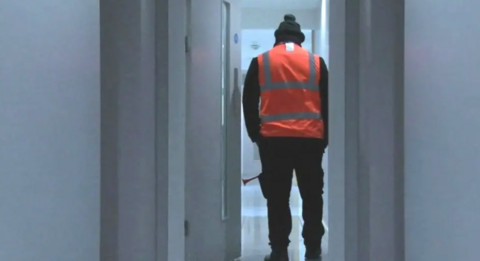 A rear view of a man wearing an orange high-vis jacket and woolly hat. He is carrying a foghorn and is standing in a doorway at the end of a white corridor.