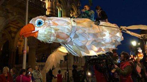 Large bird with orange beak and white and brown feathers, with white and brown wings.  It is made from paper and lighted from inside. It is being carried by various people. Lanterns are visible in a parade behind the bird.