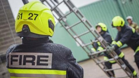 Firefighters undertaking a training exercise. One is stood with his back to the camera showing his uniform and the word "fire". In the background two firefighters are holding a large ladder against a building. 