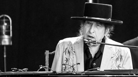 Bob Dylan, wearing a black hat and a light jacket, is seen performing on stage