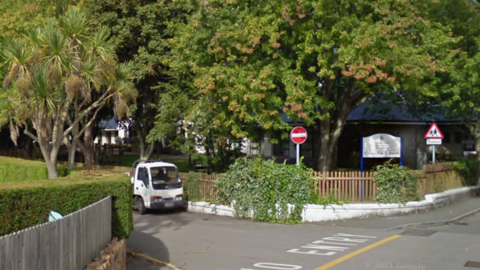 The entrance to Grouville School. There is a sign behind a wooden picket dence and there are mature trees. The road in front has NO ENTRY pianted on it and the garden opposite has a neat hedge and palm trees. There is a white van parked along the road and there are a school and no entry road signs.