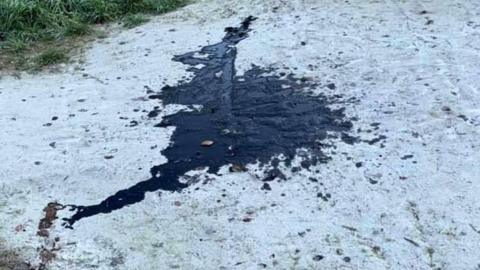 A patch of oil on a chalk ground. There oil is black in colour and rather large. There is a grass area to the left. 