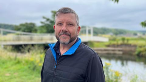 Mark Ryan wearing a dark coat, standing on a riverbank 