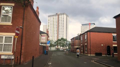 Tower blocks can be seen in the distance, behind rows of terraced red brick properties. A road sign on the left says Co-operative Street.