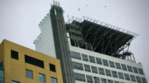 A metal platform on top of a white building, there is a yellow building to the left of it and a grey sky beyond