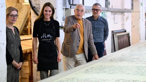 The Princess of Wales wears a black Marina Mill apron and looks toward the camera in the direction of a long printed sheet on a table. She is with three other people, two men and a woman who are explaining the item to her. 
