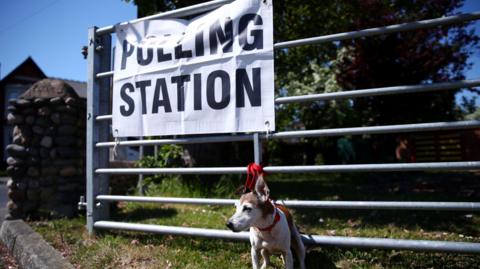 A dog tied to a gate with a red lead outside a polling station in St. Annes, Lancashire. It is a sunny day.