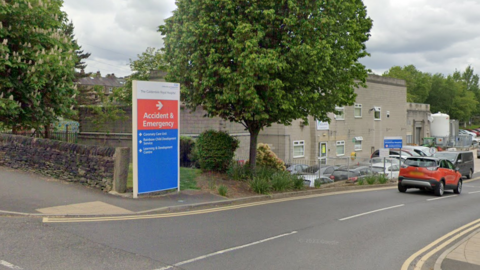 Street view shot of the welcome sign at Calderdale Royal Hospital. The sign stands on the side of the road, while a red car drives past into the car park of the hospital.