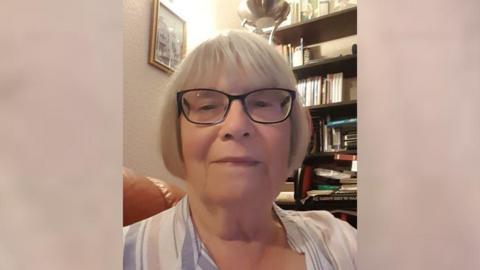 Janet Holmes with grey hair in a bob and black glasses. She is wearing a white striped shirt. She is looking at the camera. 