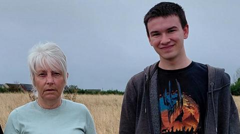 Karen Sharkey and Alex Shaw-Young pictured in a field that housing developers have earmarked for 225 new homes. They are both looking directly at the camera. Karen has short white hair and is wearing a blue t-shirt. Alex is standing next to her. He has short dark hair and is wearing a black graphic t-shirt and a black hoodie.