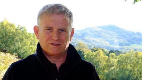 A man with grey hair is standing in front of trees and a hill. He is wearing a dark top.