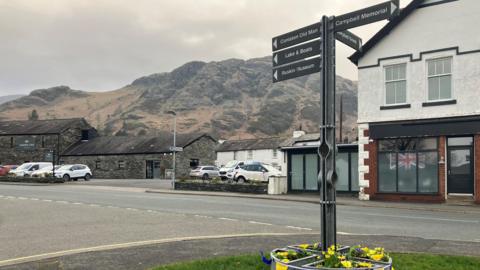 A general view of the village of Coniston. There is a sign in the foreground signposting to various attractions in the village. There are shops and buildings lining the main street and the fells in the background.