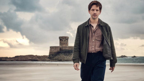 Damian Moloney as Jim Bergerac on a beach in Jersey. He is wearing a jacket and a button up shirt and black trousers. The man has brown hair and a facial hair