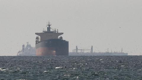 Tanker sails on open sea in the Gulf near the Strait of Hormuz, while several other ships are silhouetted in the background behind it