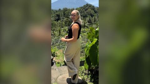 A woman wearing a back sleeveless top and beige trousers stands with a forest in the background