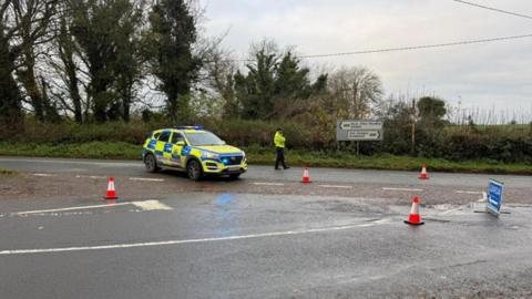A Garda patrol car is parked at a road junction with a Garda officer standing nearby. There are traffic cones on the road and diversion sign.