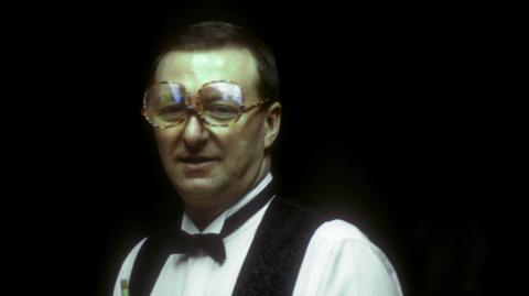 Dennis Taylor wearing a white shirt, black waistcoat and black bow tie and his trademark large glasses.