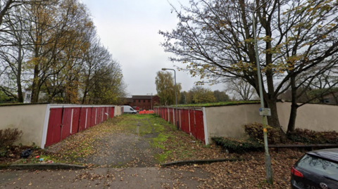 The garages off Zoar Street, Wolverhampton, which are set to be demolished. They have red doors and are pictured on the left and right of the image with a grassy concrete section in between them. Leaves have fallen on the ground and trees stand behind them as well as lamp posts.