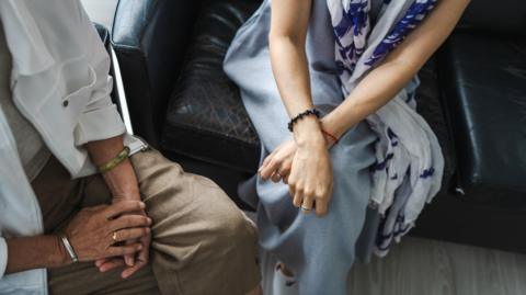 Two people, a man and a woman, sit in a counselling session, their hands folded in their laps. Their faces are out of shot.