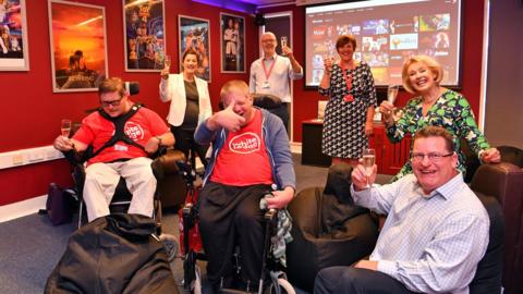 Service users at The White Lodge Centre in Chertsey, Surrey, alongside employees of the company pictured after a cinema room was installed. Staff are all holding up a glass of fizz and there are two service users in wheelchairs.