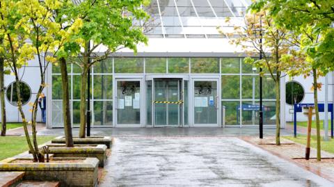 File image from 2015 showing the walkway and entrance at Woolwich Crown Court. The building has a glass facade with three glass doors in the centre - the centre is a revolving door. The path towards the court is lined with small trees.