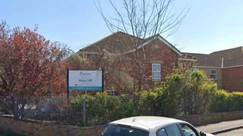 Google Street View of Muscliff Nursing Home a large, modern, two-storey red brick building with a pitched roof behind a hedge and railings.