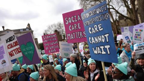 A crowd of people, many wearing teal coloured hats hold placards with the phrases: "#ProvidersUniteStronger Together - care cannot wait" and "Social Care Matters"