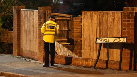 A police officer looking at a destroyed section of fencing.  A road sign reading 'Church Avenue' is next to the fence and has also been damaged.