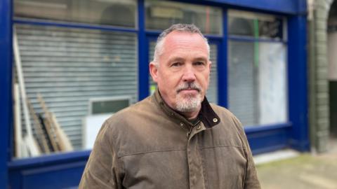 A man in a brown coat stands out a shuttered shop in Pontefract. The shopfront is blue