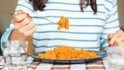 A person sitting at a table eating penne pasta from a plate with a fork.
