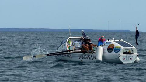 A mid-shot of two people rowing away from the camera in a boat covered with stickers