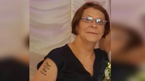 Carol-Ann Rimini, looking at the camera, she is wearing glasses, has short brown hair and is wearing a sleeveless black top with a necklace. She is standing in front of a wall with white  fabric on it and has a tattoo on her right shoulder. 